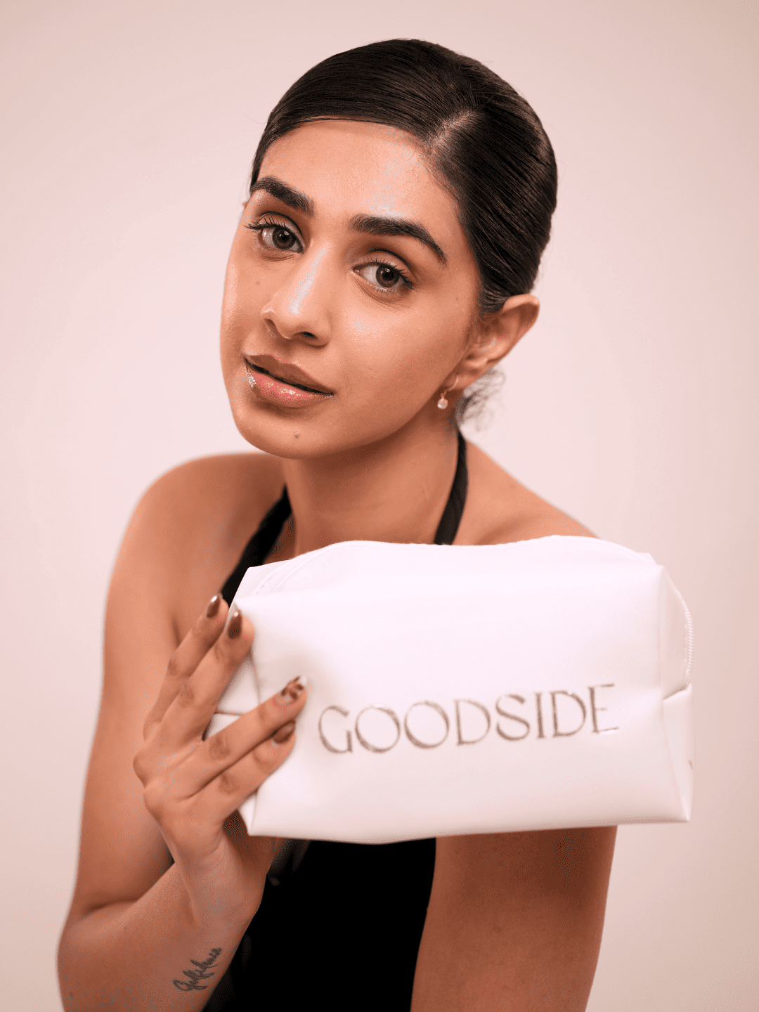 A woman with dark hair pulled back holds a white cosmetic pouch with the brand name 'GOODSIDE' printed in gold lettering. She is wearing a black halter top against a solid light beige background. - GoodSide
