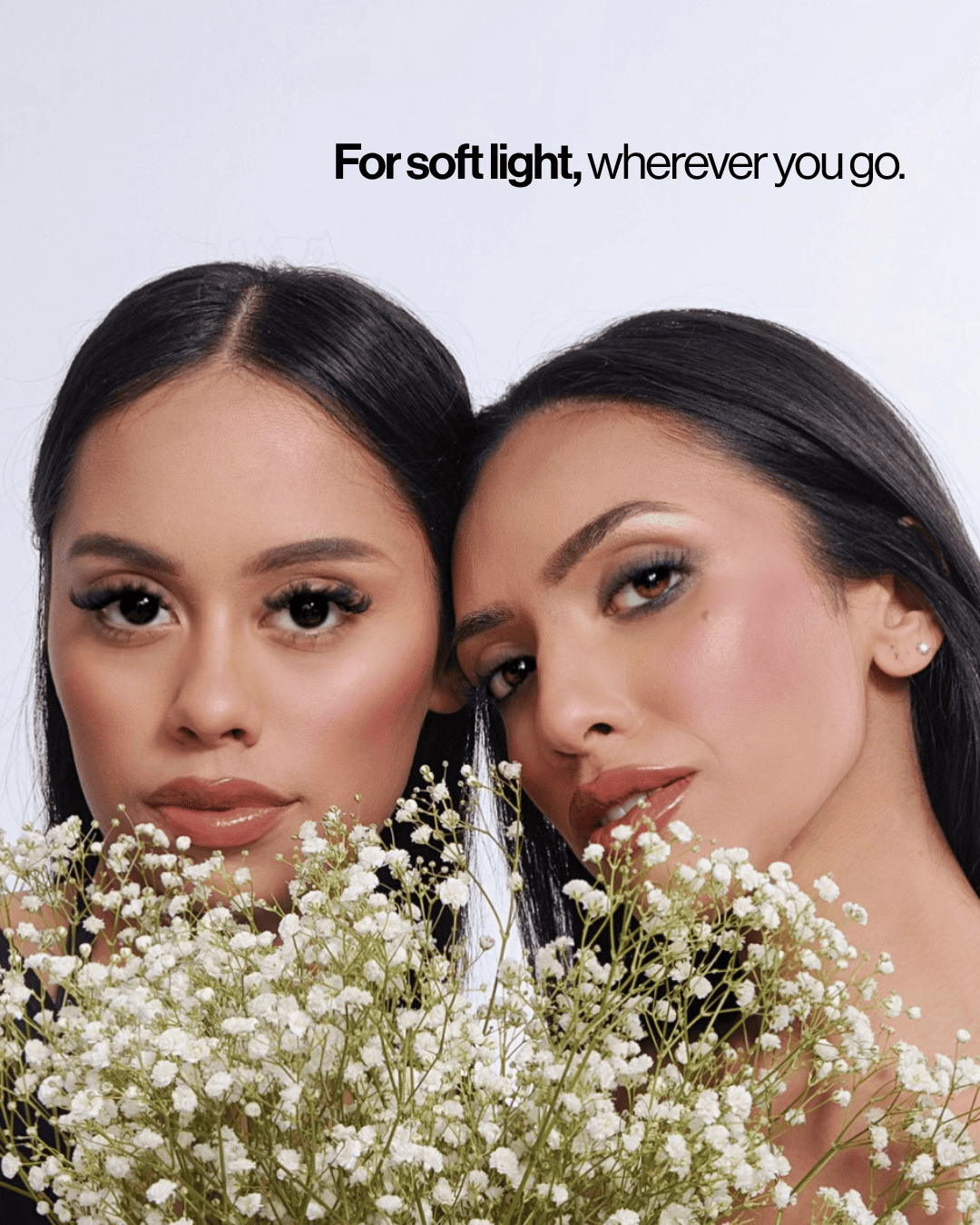 Two women with dark hair and makeup pose closely together behind a large bouquet of white baby's breath flowers, with the text 'For soft light, wherever you go.' at the top against a light gray background. - GoodSide