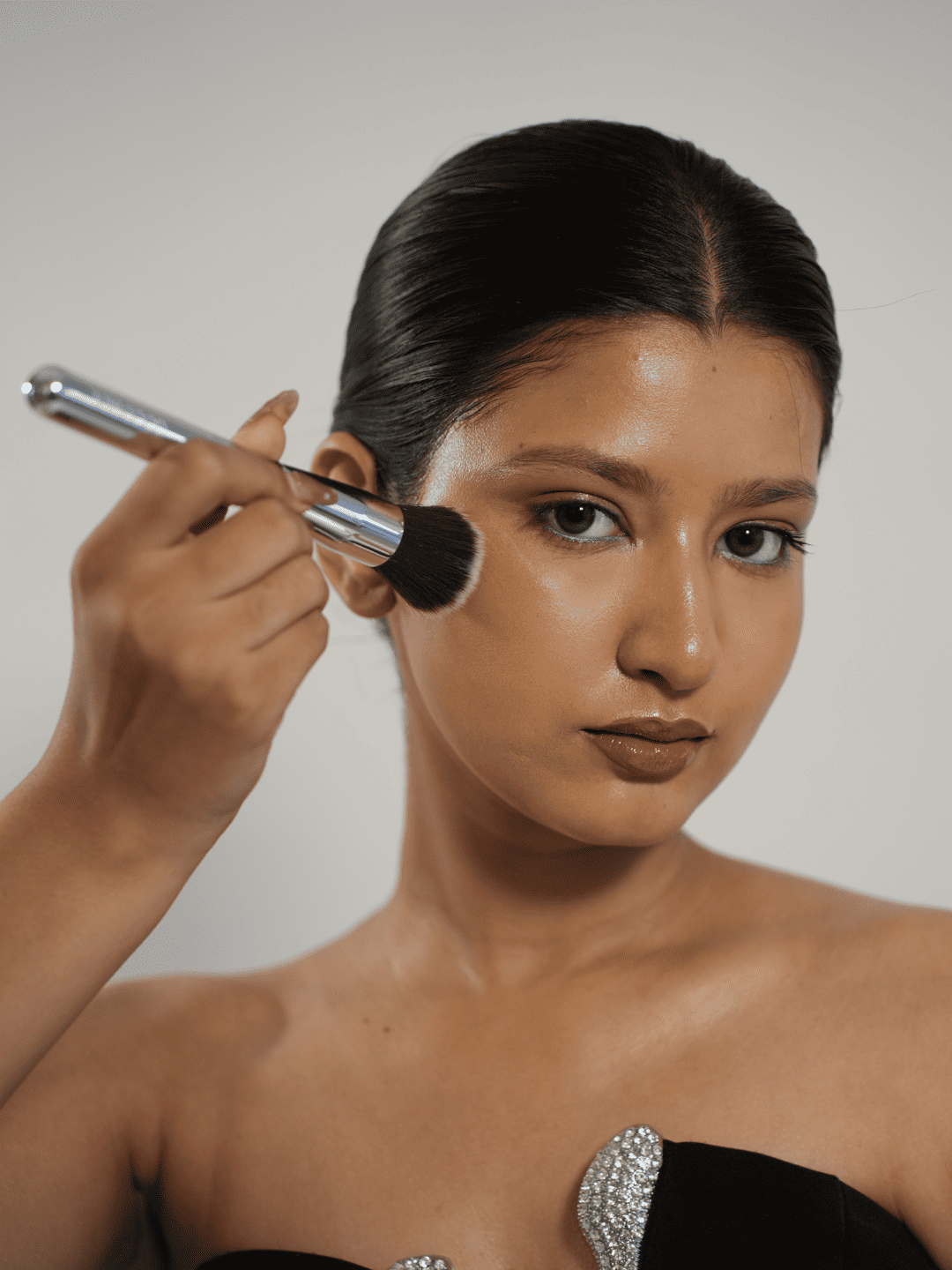 A woman with slicked-back dark hair applies blush makeup using a makeup brush, wearing a black strapless top with a rhinestone embellishment against a plain light gray background. - GoodSide