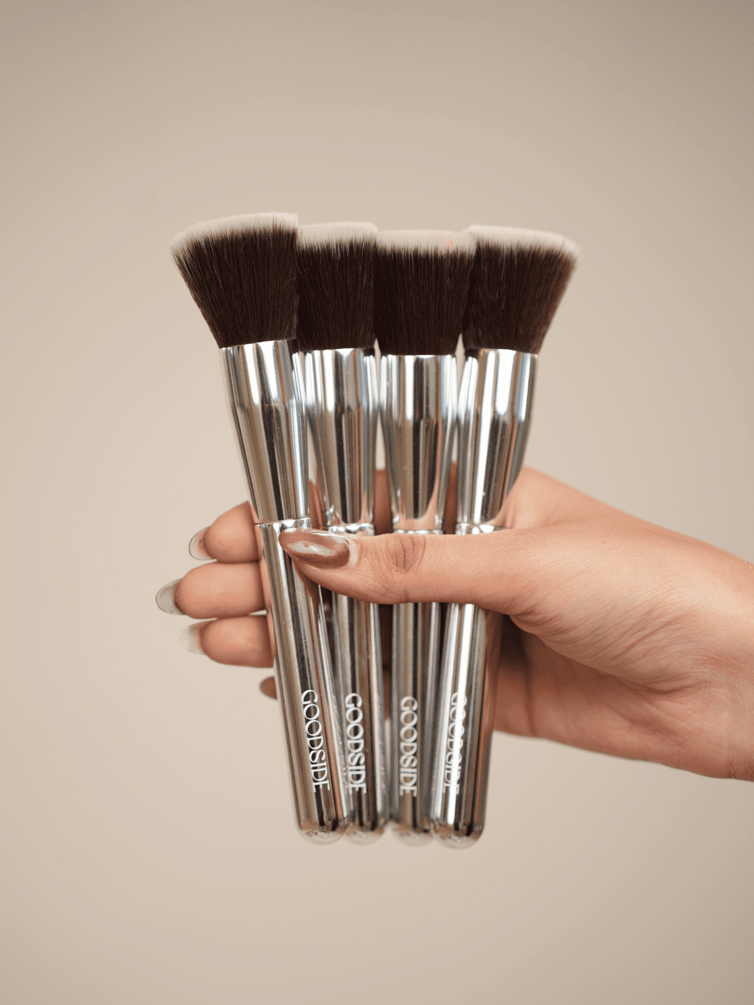 A hand holding a set of four COSISET makeup brushes with silver handles and dark brown bristles against a solid beige background. - GoodSide